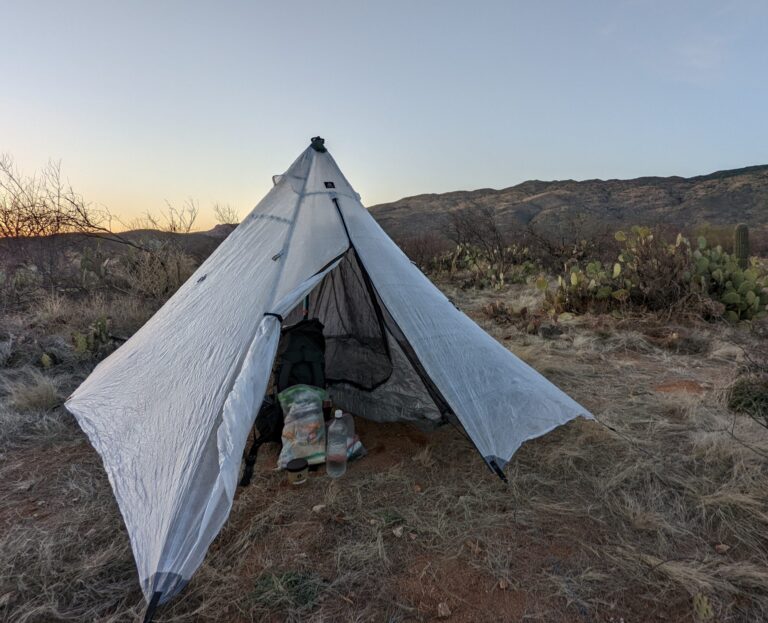 A small pyramid tarp tent at sunset on a grassy ridge, supported by a single trekking pole with stakes and guylines.