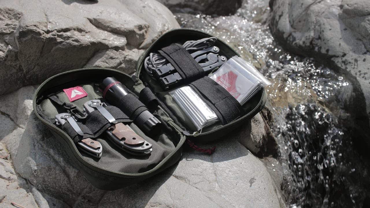 Compact survival kit with knife, flashlight, multitool, and emergency gear laid out on rocks beside a stream for outdoor survival.