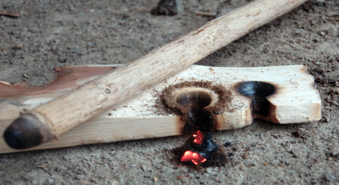 Survivalist saws a bow‑drill spindle against a cedar fireboard; thin smoke rises as friction forms a glowing ember.