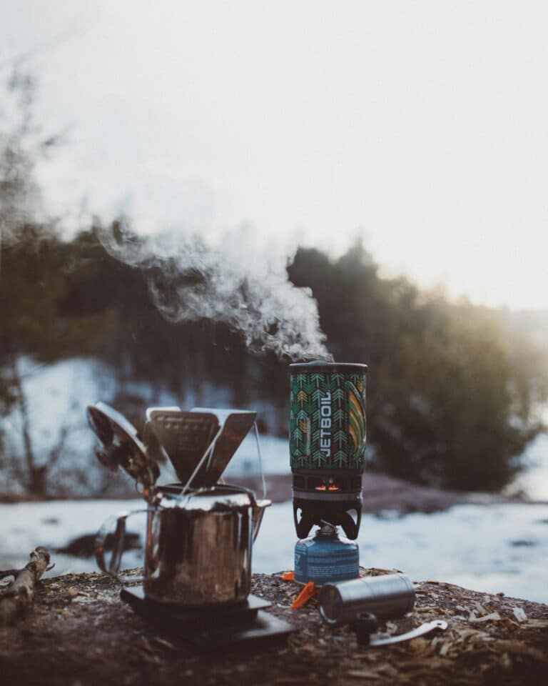 Camping stove boiling water in a Jetboil system with steam rising, next to a metal pot and lighter on a snowy outdoor campsite