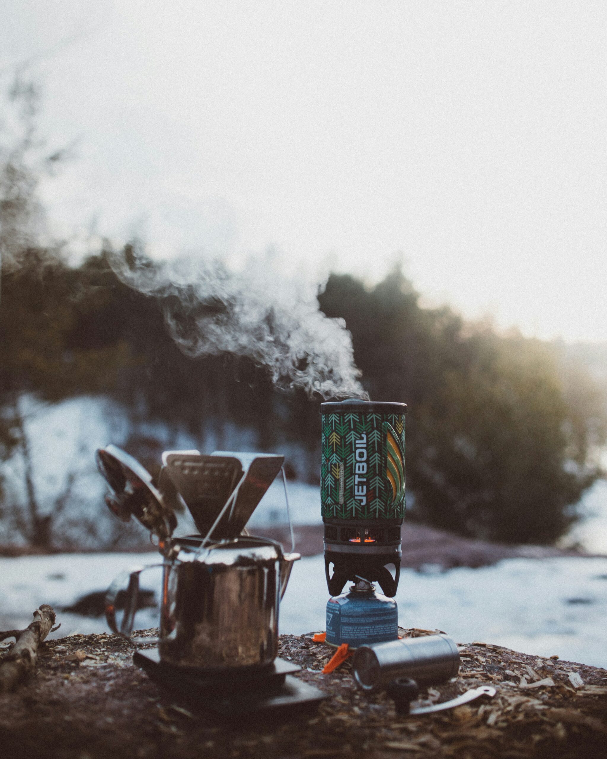Camping stove boiling water in a Jetboil system with steam rising, next to a metal pot and lighter on a snowy outdoor campsite