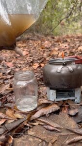 Homemade gravity water filter made from plastic and cloth pouring into glass jar in a woodland bushcraft camp
