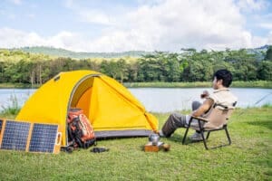 Man camping beside yellow tent with portable solar panels and backpack near a lake, enjoying outdoor survival lifestyle