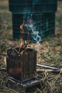 Portable survival stove burning twigs with smoke and flame, used as a fire starter in an outdoor wilderness setting