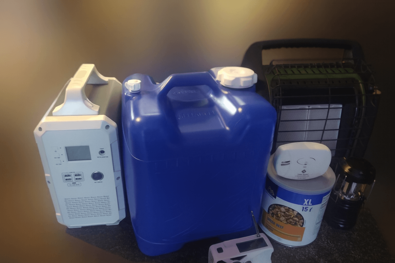 Essential emergency preparedness gear arranged on a countertop, including a portable power station, blue water container, propane heater, canned food, carbon monoxide alarm, radio, and lantern, with a softly blurred background to highlight the products.