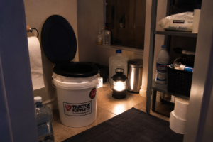 Emergency blackout sanitation setup inside a home bathroom with a bucket toilet, lantern lighting, water storage containers, hygiene supplies, and emergency cleaning products