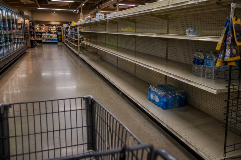 Empty grocery store shelves with only a few cases of bottled water remaining during an emergency supply shortage