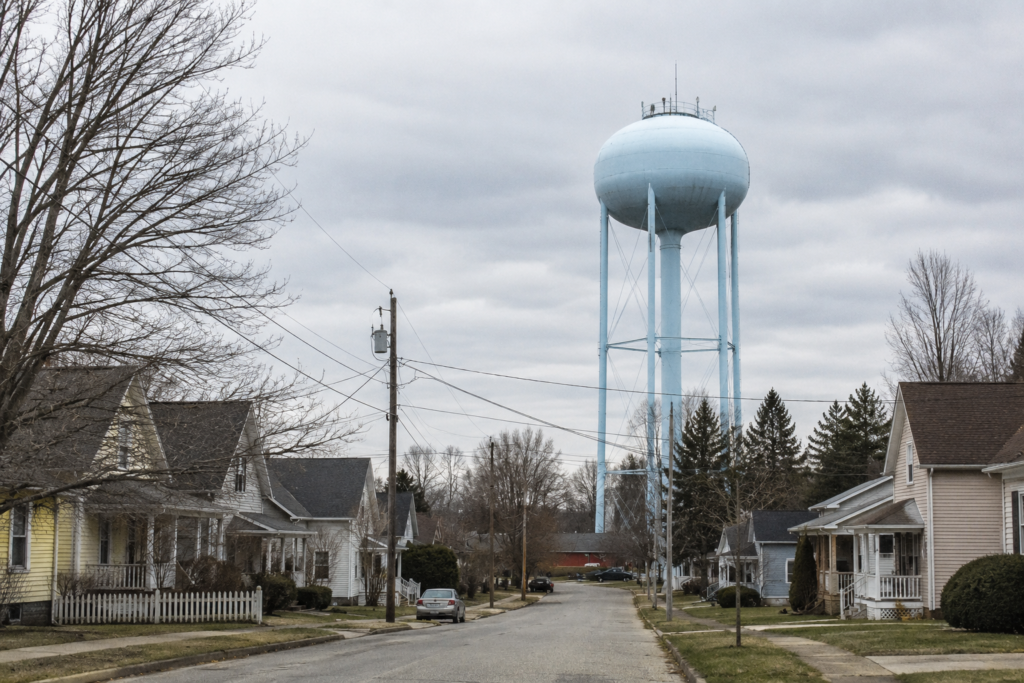 Residential neighborhood with a large municipal water tower under an overcast sky, illustrating city water infrastructure and how municipal water systems may operate during a prolonged power outage or blackout.
