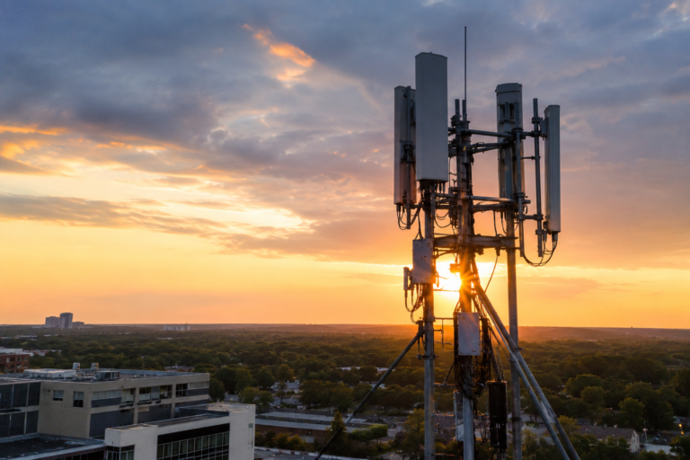 Cell tower antennas operating above a city skyline at sunset, illustrating cellular communication infrastructure and backup network systems during a power outage or blackout.