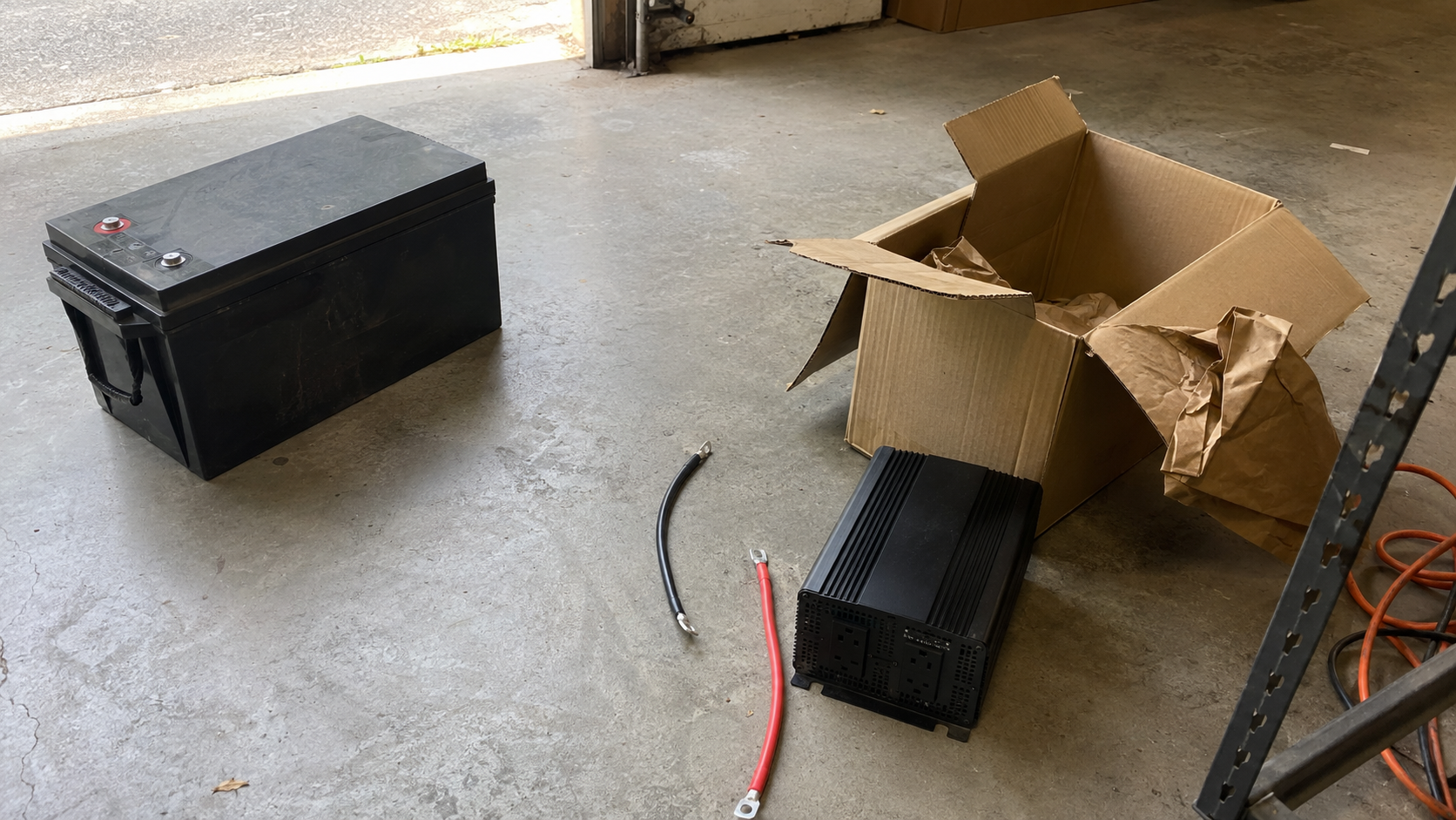 12V 200Ah battery beside a pure sine wave inverter, battery cables, and an open box on a garage floor.