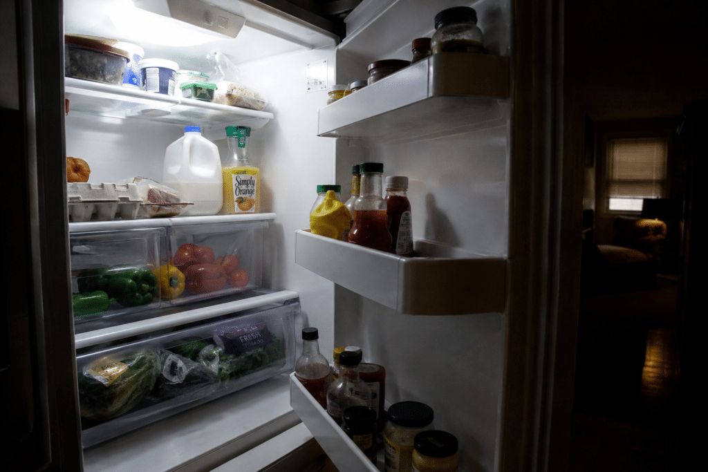 Open household refrigerator filled with fresh food, milk, eggs, vegetables, leftovers, and condiments during a nighttime power outage, illustrating food safety and cold storage preparedness.