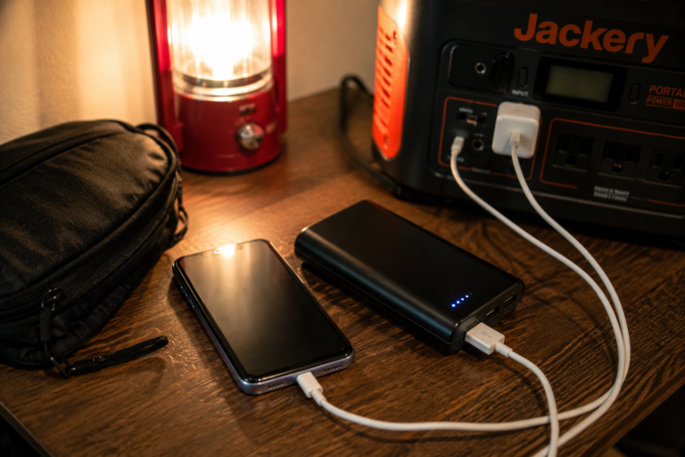Smartphone charging from a portable power bank and portable power station beside a lit emergency lantern during a home power outage, demonstrating reliable blackout backup charging solutions.